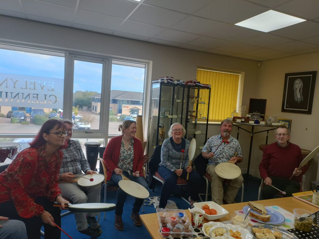 Image Description: A set of five photographs, showing Evelyn and members of the Volunteer team. All of the photos show a variety of the Volunteer team smiling, some with people sitting and playing Uchiwa Daikos and Remo Paddle Drums, and some photos with people stood casually chatting with a light buffet on a table in the middle of the room.