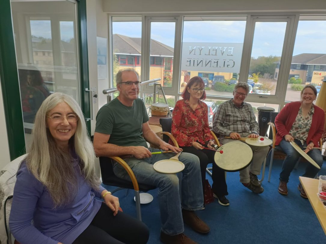 Image Description: A set of five photographs, showing Evelyn and members of the Volunteer team. All of the photos show a variety of the Volunteer team smiling, some with people sitting and playing Uchiwa Daikos and Remo Paddle Drums, and some photos with people stood casually chatting with a light buffet on a table in the middle of the room.