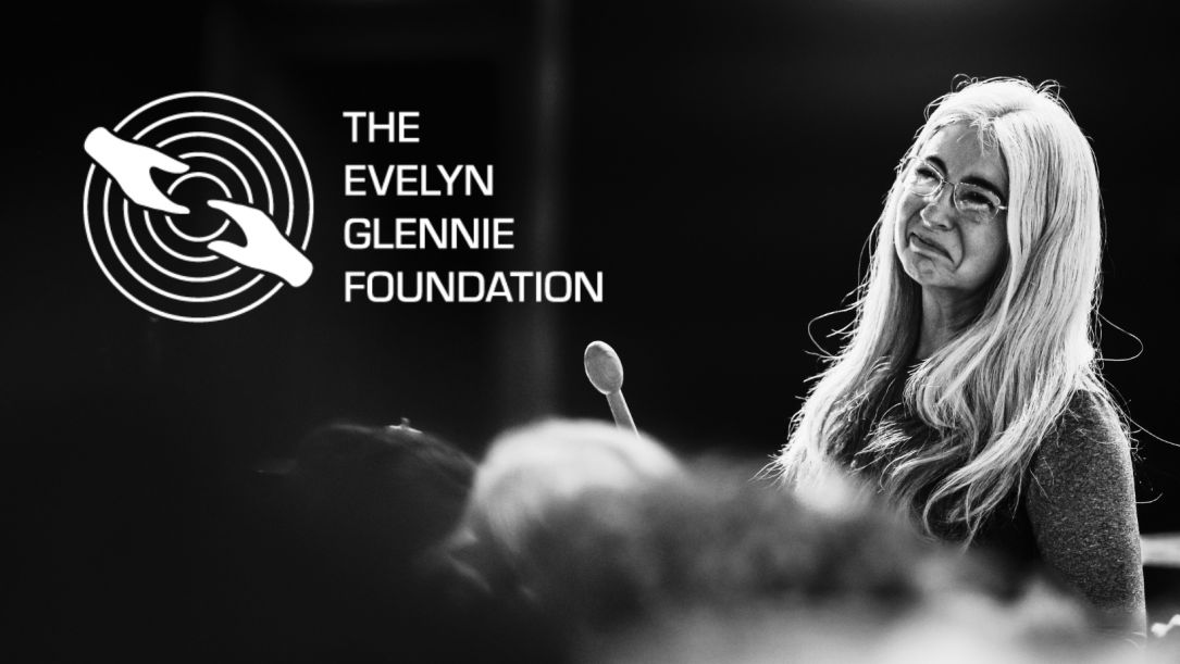 Image description: A black and white photo of Evelyn, with The Evelyn Glennie Foundation logo in white floating over a dark section. The logo is a series of five concentric circles, with two hands reaching to meet each other, one of which is helping the other 'up'. In the photo, Evelyn is wearing her glasses during a conversation, and is smiling broadly.